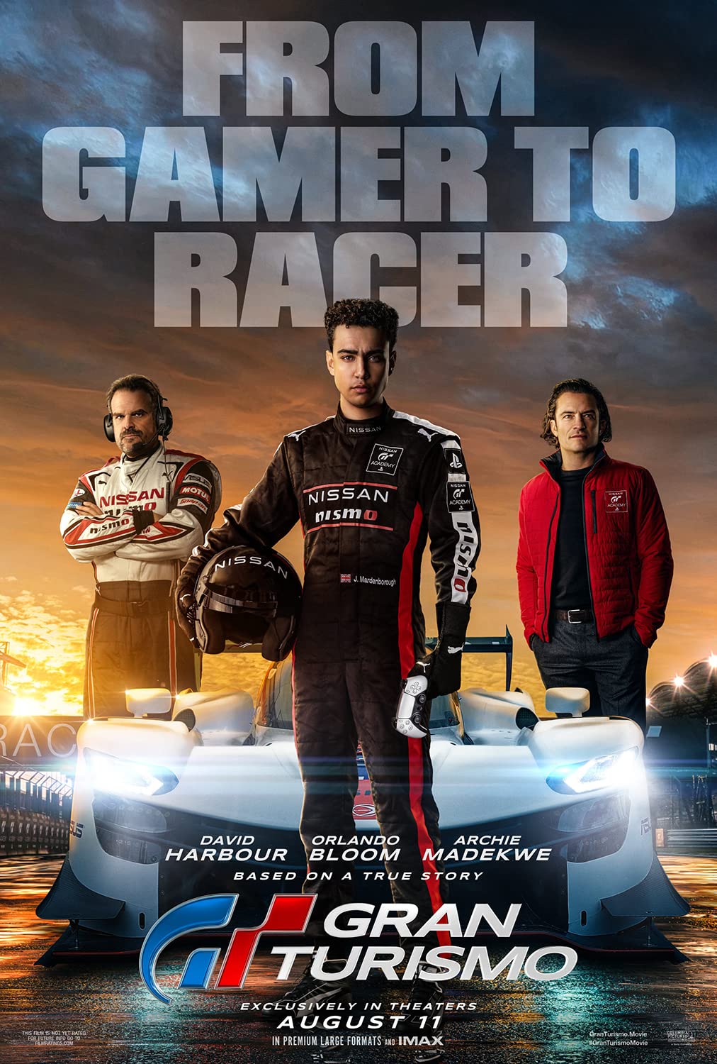The movie poster for Gran Turismo. Two middle aged men and a boy in his late teens stand around a race car. Text at the top of the poster reads 'From Gamer to Racer'